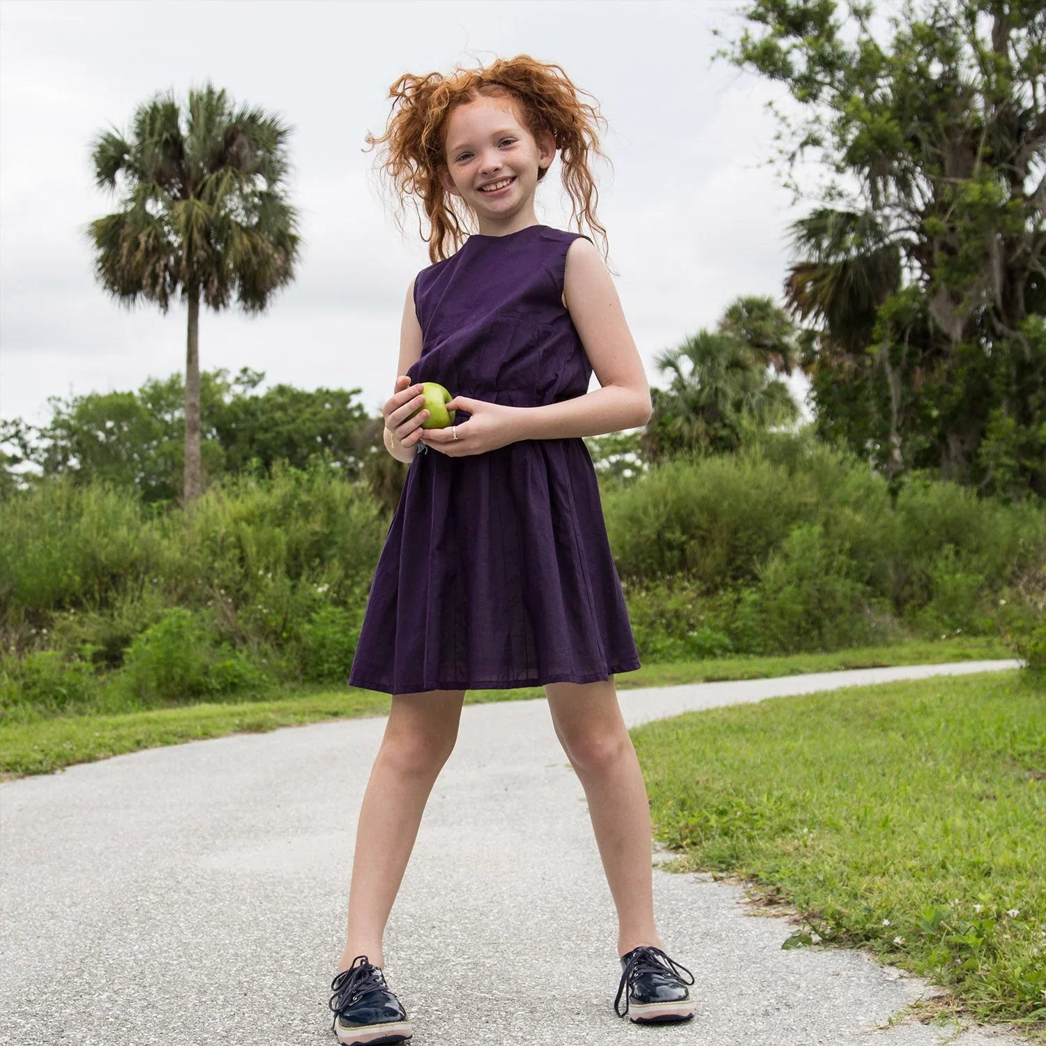 Cute girl wearing a Girls Beach Dress in Purple, holding an apple, posing outdoors on a sunny day.