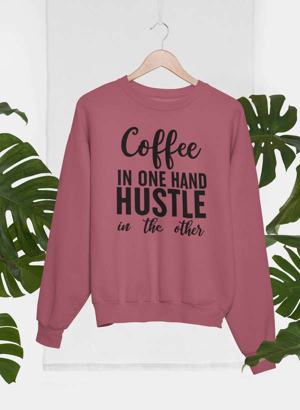 Coffee In One Hand Hustle In The Other Sweat Shirt displayed on a hanger against green leaves.