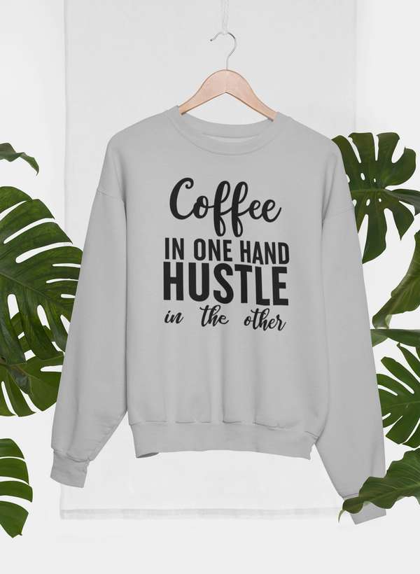 Coffee In One Hand Hustle In The Other Sweat Shirt displayed among green leaves on a hanger.